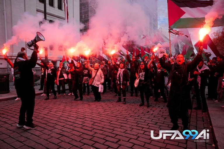 กลุ่มผู้ชุมนุมถือธงปาเลสไตน์และจุดพลุควันสีแดงระหว่างการเดินขบวนประท้วงกลางเมือง ท่ามกลางบรรยากาศตึงเครียดและแสงควันปกคลุมทั่วบริเวณ ภาพของ ufa99ai